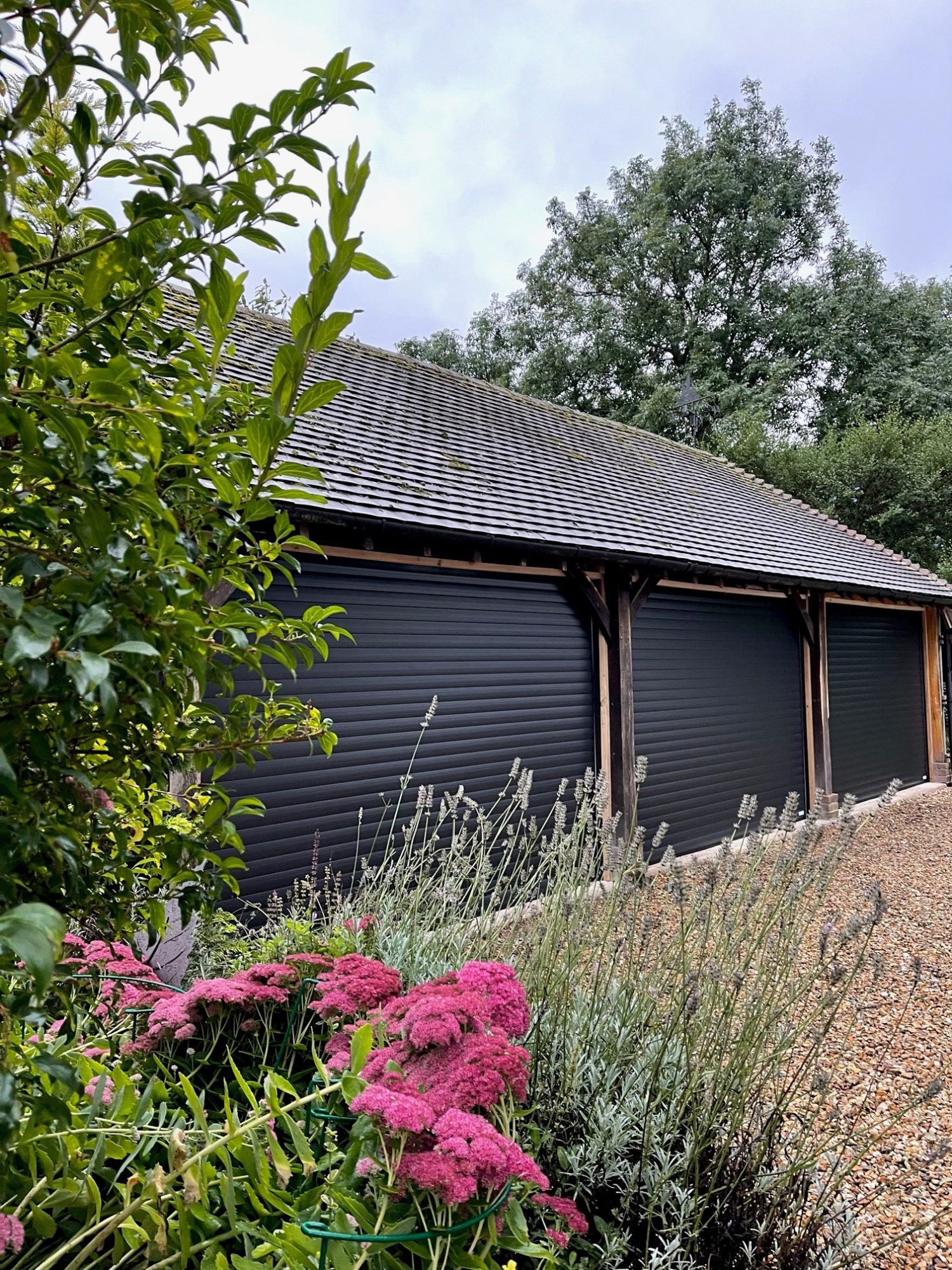 Detached garage building with three separate garage doors featuring black roller doors and summer flowers in the foreground.